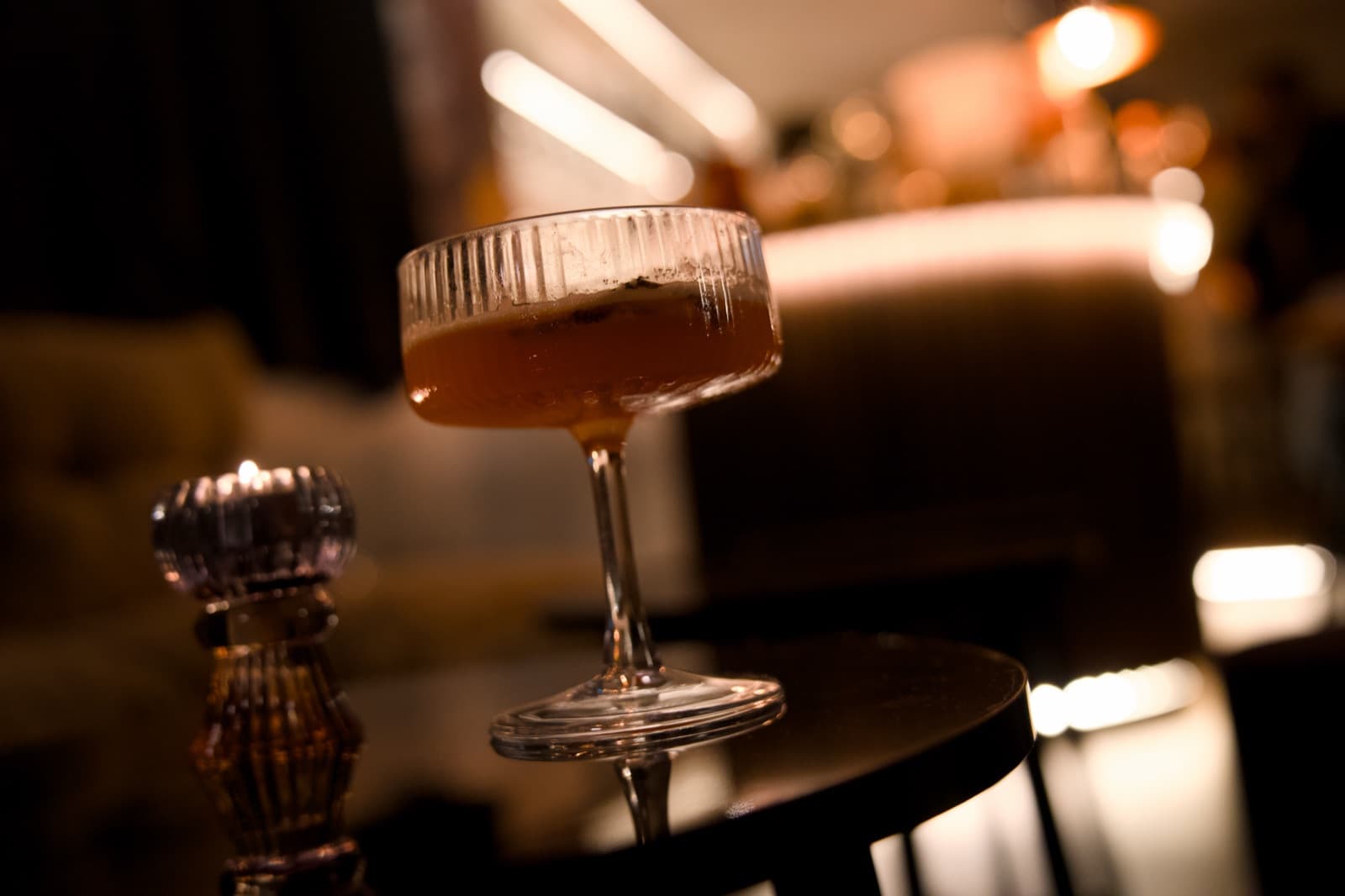 A coupe cocktail on a low table, the bar glowing behind