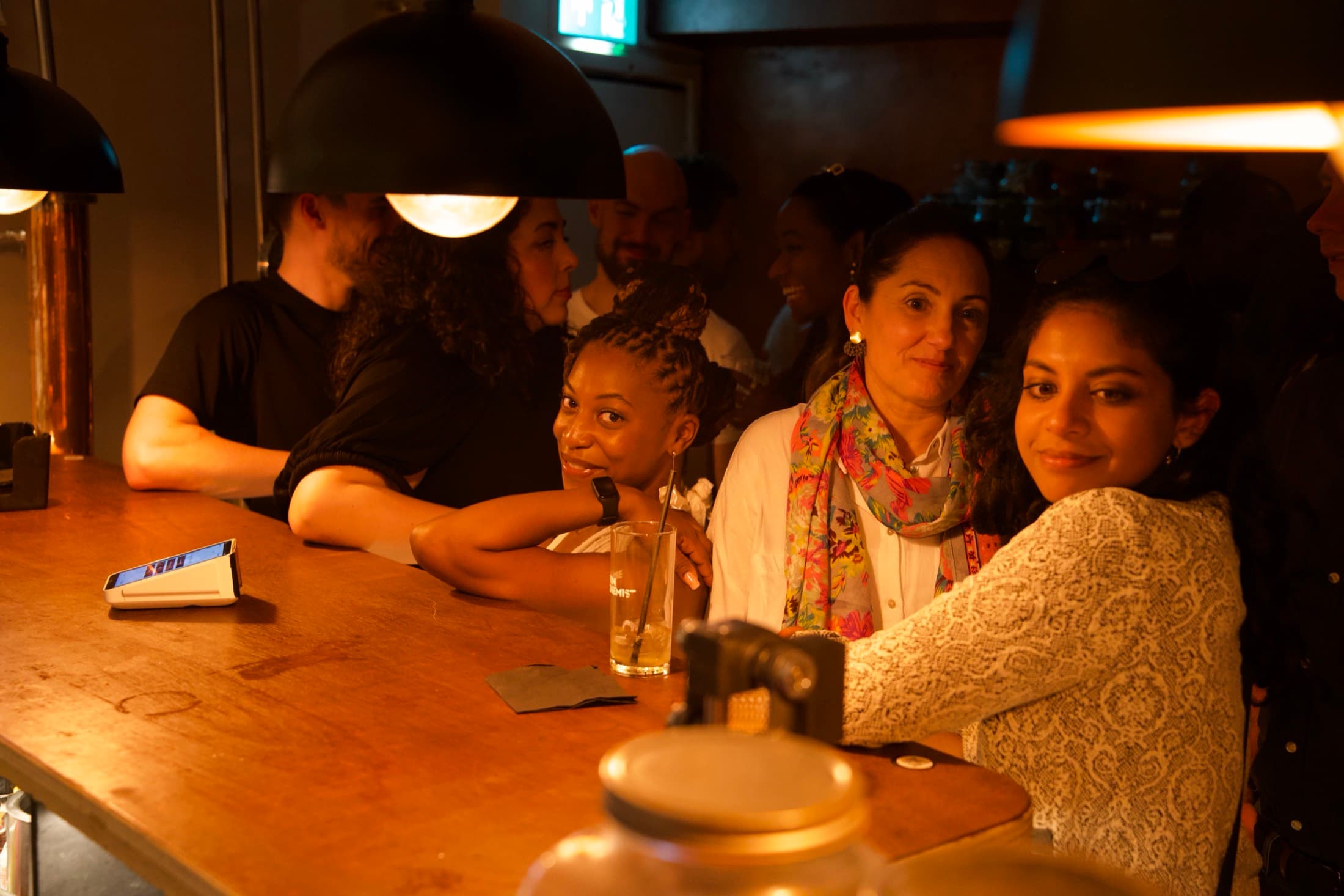 A group leaning on the bar together, drinks in hand, pendant lights above