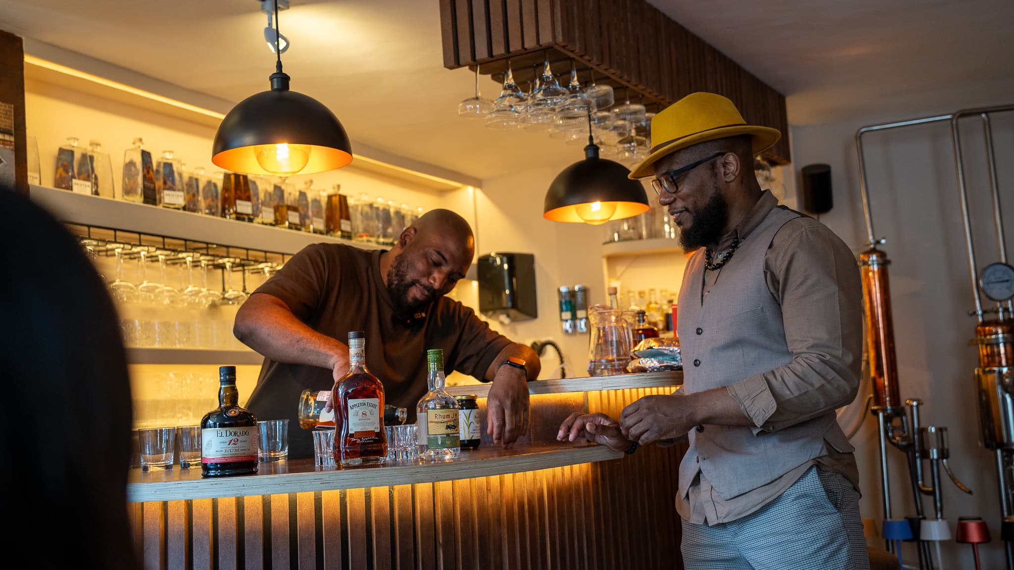 Two hosts behind the bar, one pouring, the house bottles lined along the shelf above