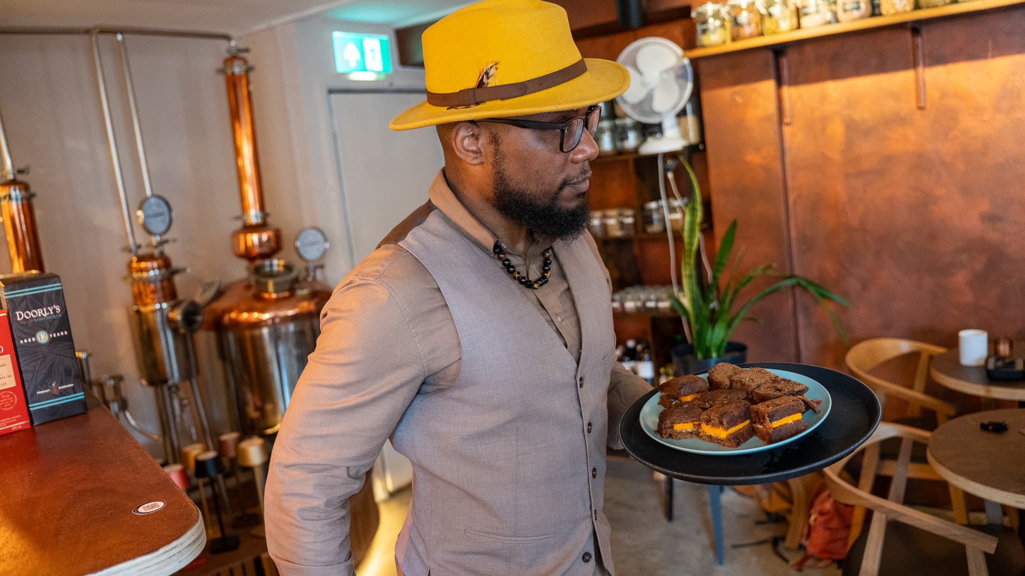 A host carrying a tray of Caribbean snacks past the copper stills